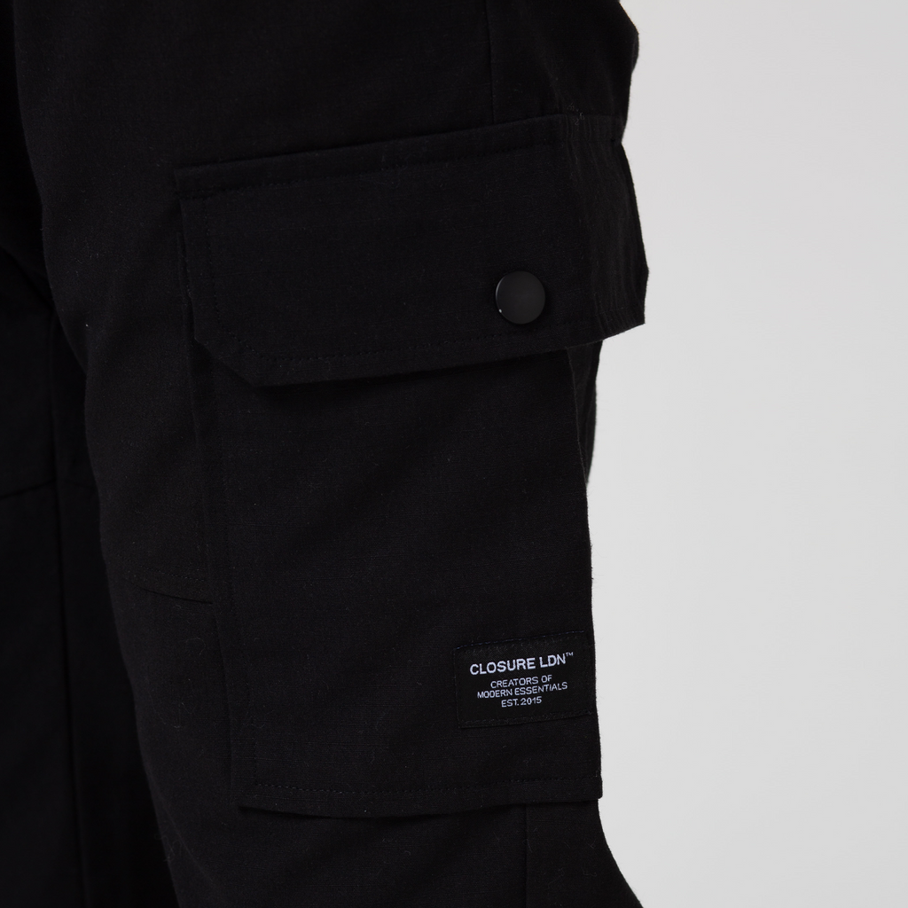 Close up of black cargo pants pocket with black popper and black logo with white writing "CLOSURE LDN"
