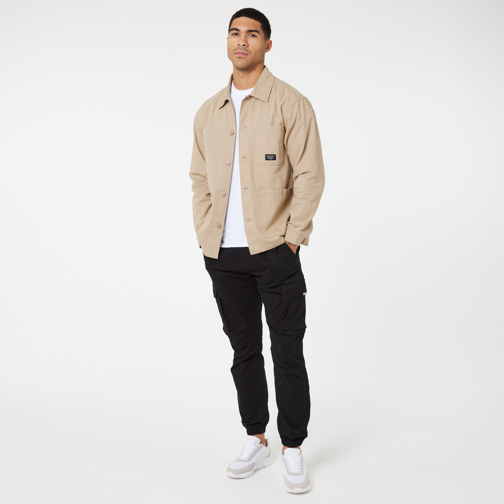 Model wearing men's overshrit jacket in stone unbuttoned with white tee and black cargo pants