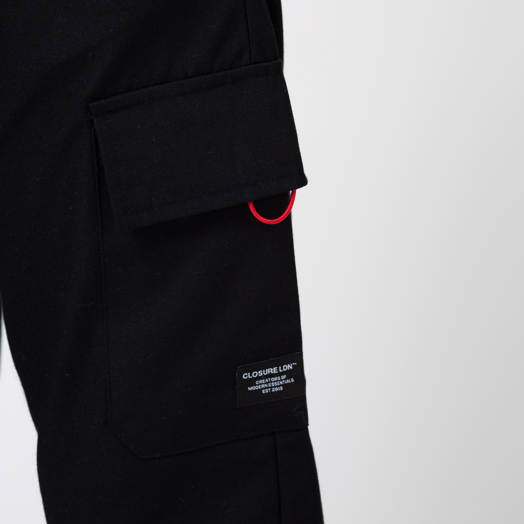 Close up image of black utility cargo pants pocket with red tag and black patch with logo in white writing