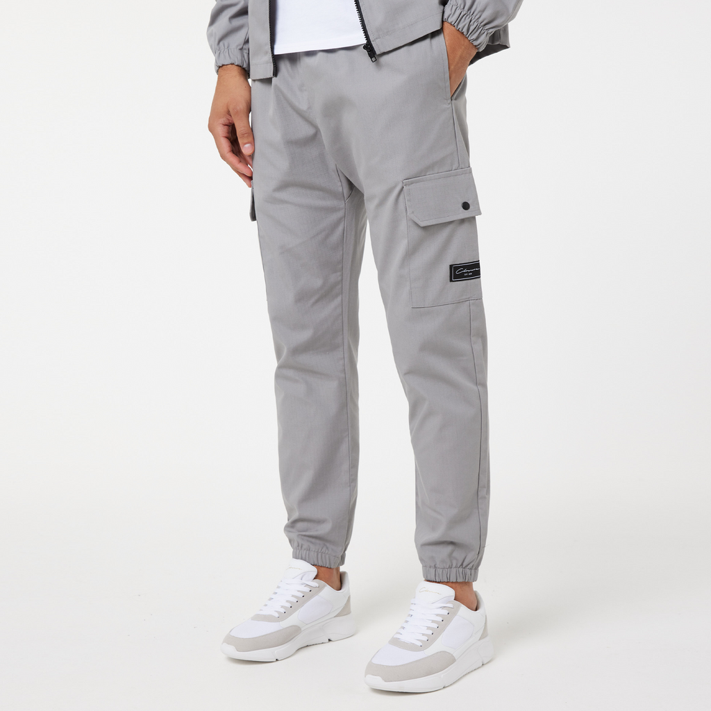 model wearing men's utility trousers in grey with a black square logo and styled with white casual trainers