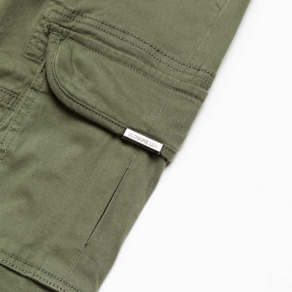 Extreme close up on green utility trousers pocket with silver metal tag on pocket lid saying "Closure LDN"