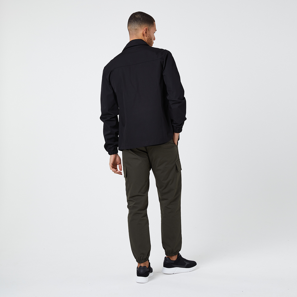 Back profile view of model wearing dark green cuffed cargo pants with trainers and black overshirt