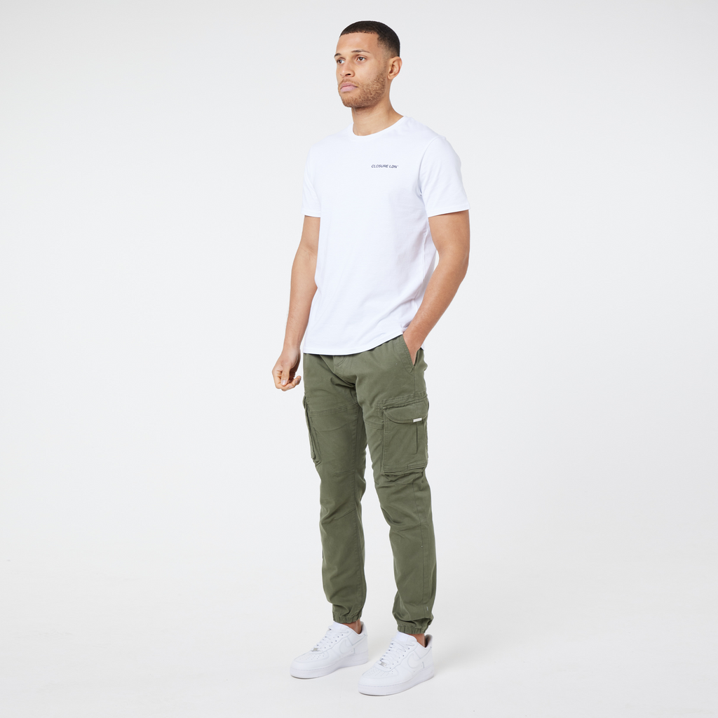 Model showing full outfit of kahki green utility trousers paired with a mens white top with a small black logo and trainers